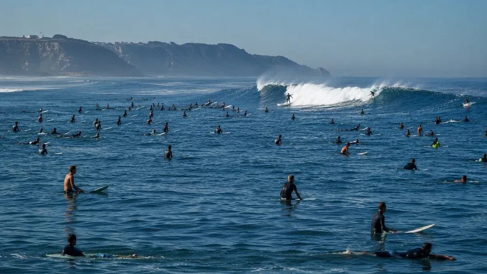 Dezenas de surfistas sentados nas pranchas no outside esperando a série, com dois surfistas descendo a mesma onda ao fundo