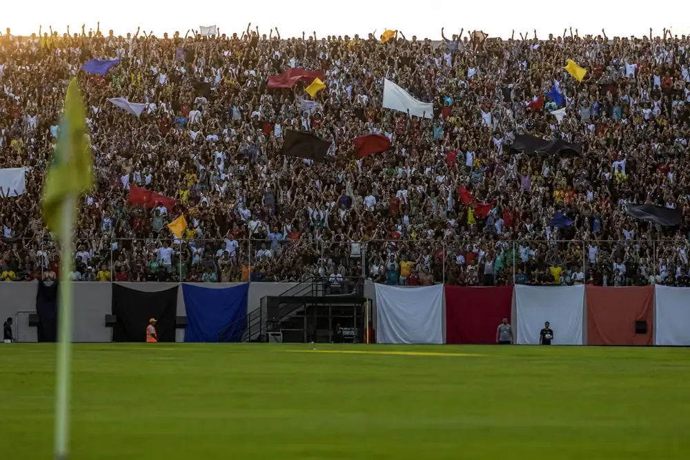 Arquibancada de estadio de futebol brasileiro lotada, com torcedores de pe comemorando e bandeiras lisas sem escudos